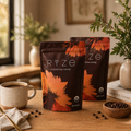 Two bags of RYZE Mushroom Coffee standing on a wooden table in a warm, styled interior. A ceramic mug of coffee, scattered coffee beans, books, and potted plants are arranged around the packages.