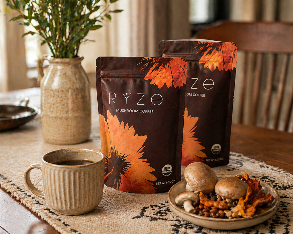 Two bags of RYZE Mushroom Coffee standing on a table runner beside a ceramic mug of coffee and a plate of nuts and dried fruit, with a vase of greenery in the background.