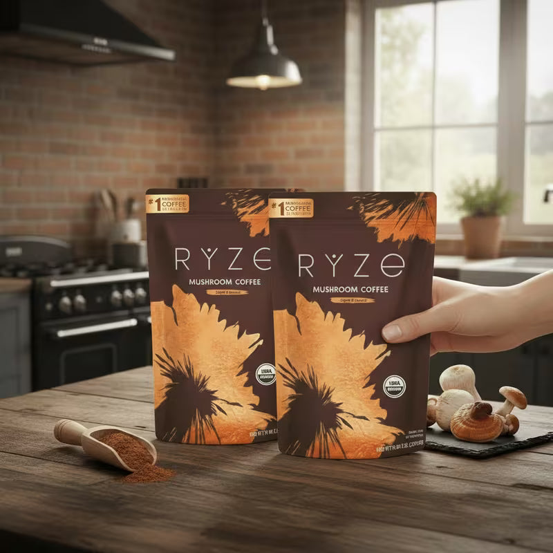 Two bags of RYZO Mushroom Coffee standing on a rustic wooden table in a kitchen setting, with a hand holding one bag. A wooden scoop of coffee powder and whole mushrooms are placed nearby.