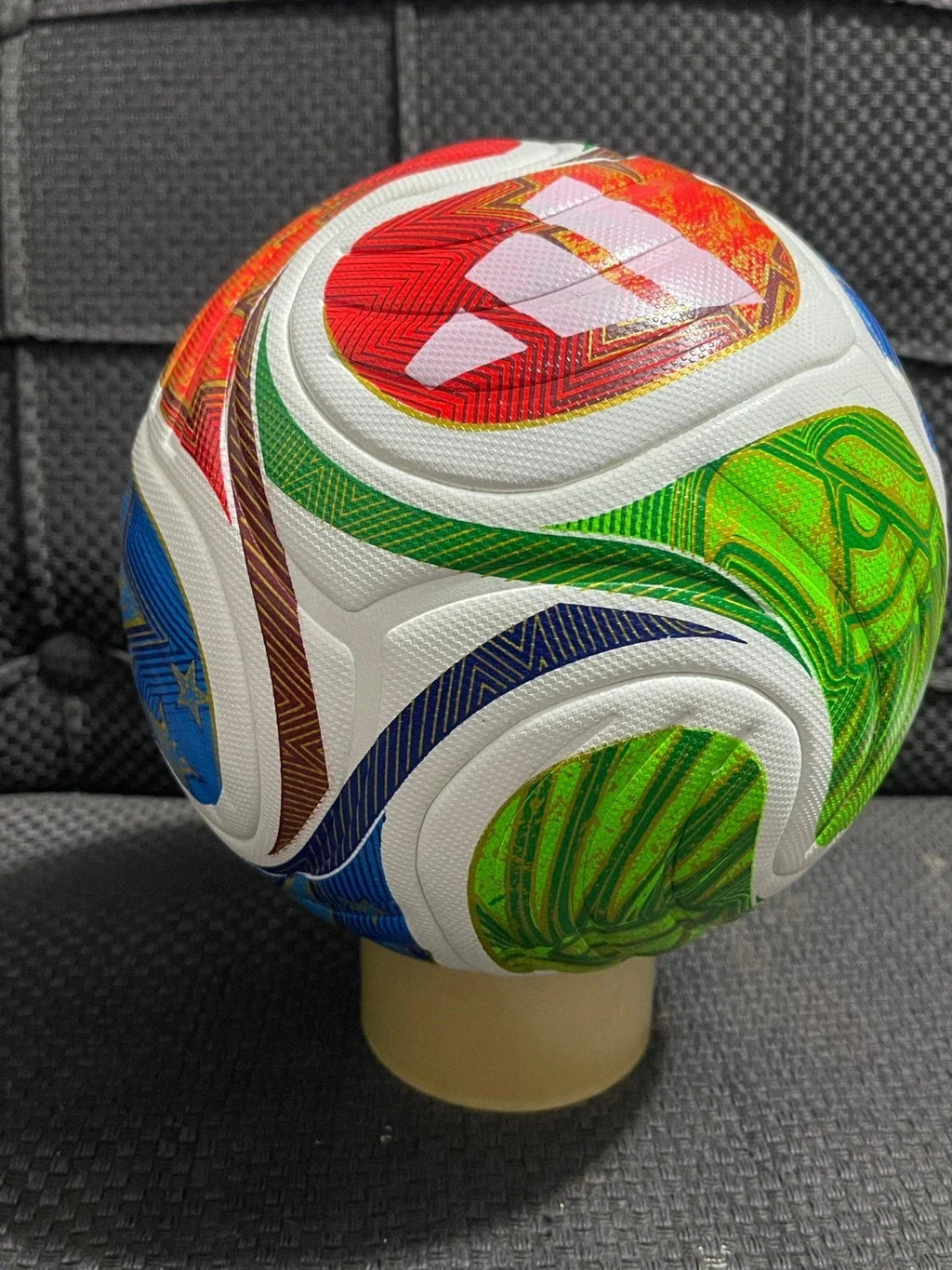 A colorful Adidas soccer ball with white, green, blue, red, and brown panels, displayed on a beige plastic stand against a dark textured background.