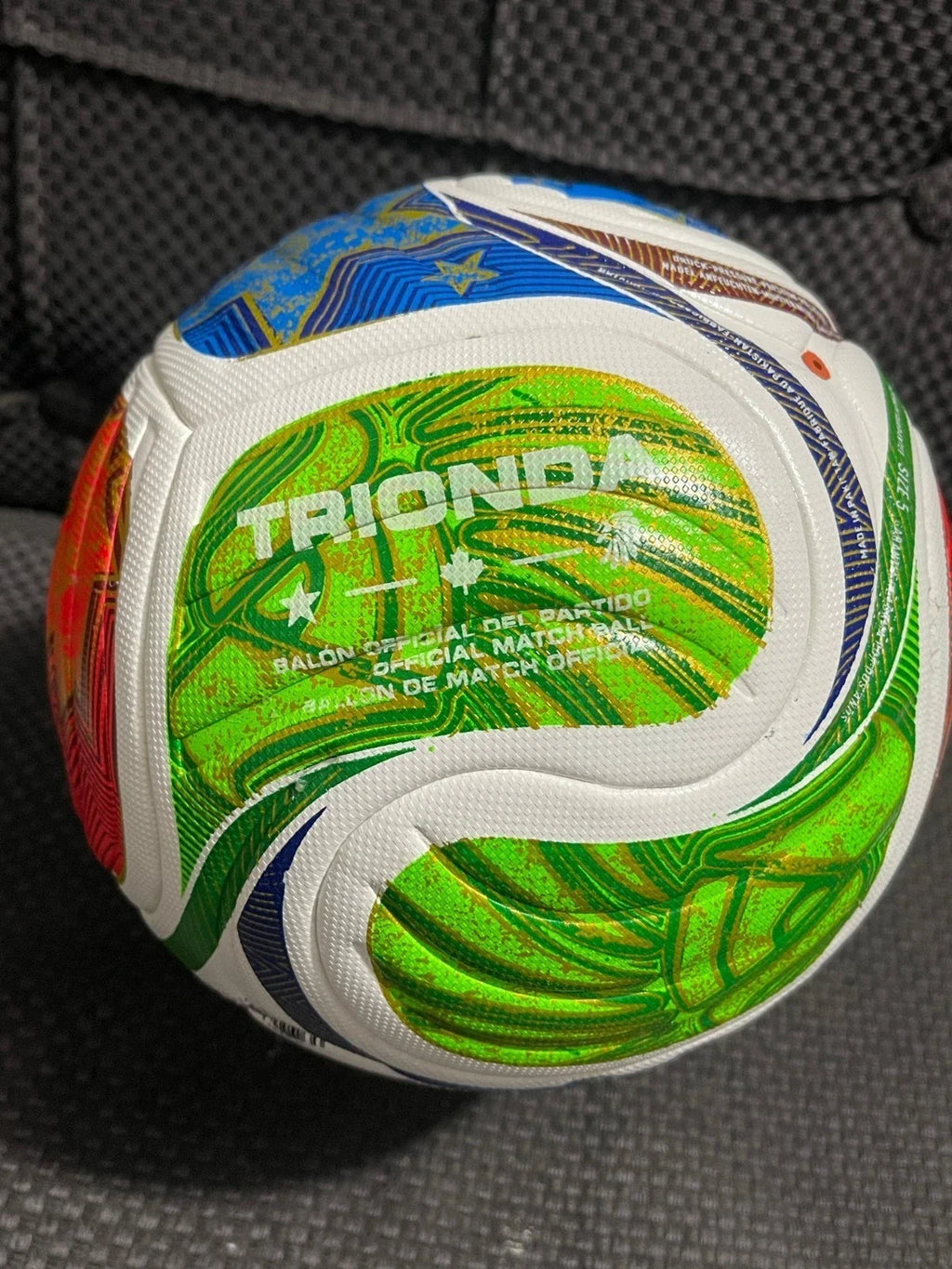 A Trionda soccer ball with a green, white, and blue leaf pattern, resting on a black textured fabric surface.