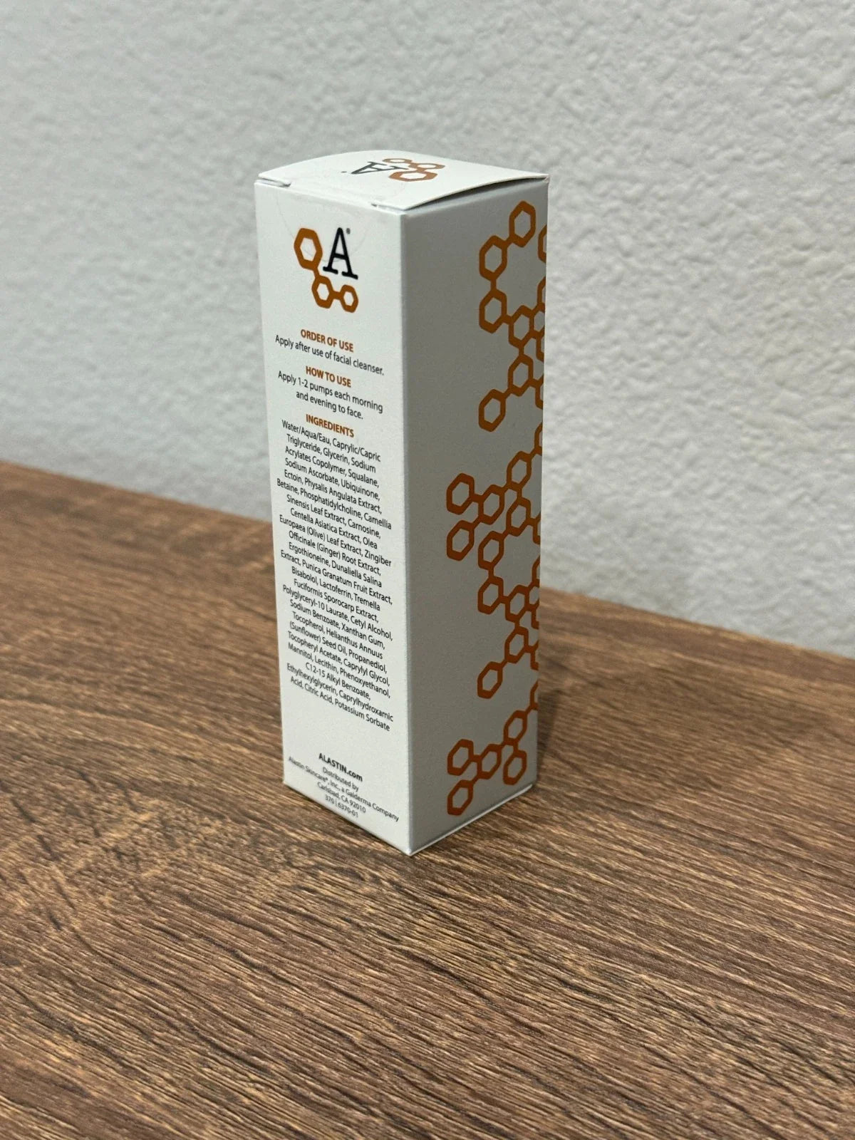 A tall white rectangular product box with orange circular honeycomb-style graphics and printed text, standing on a wooden tabletop against a light textured wall.