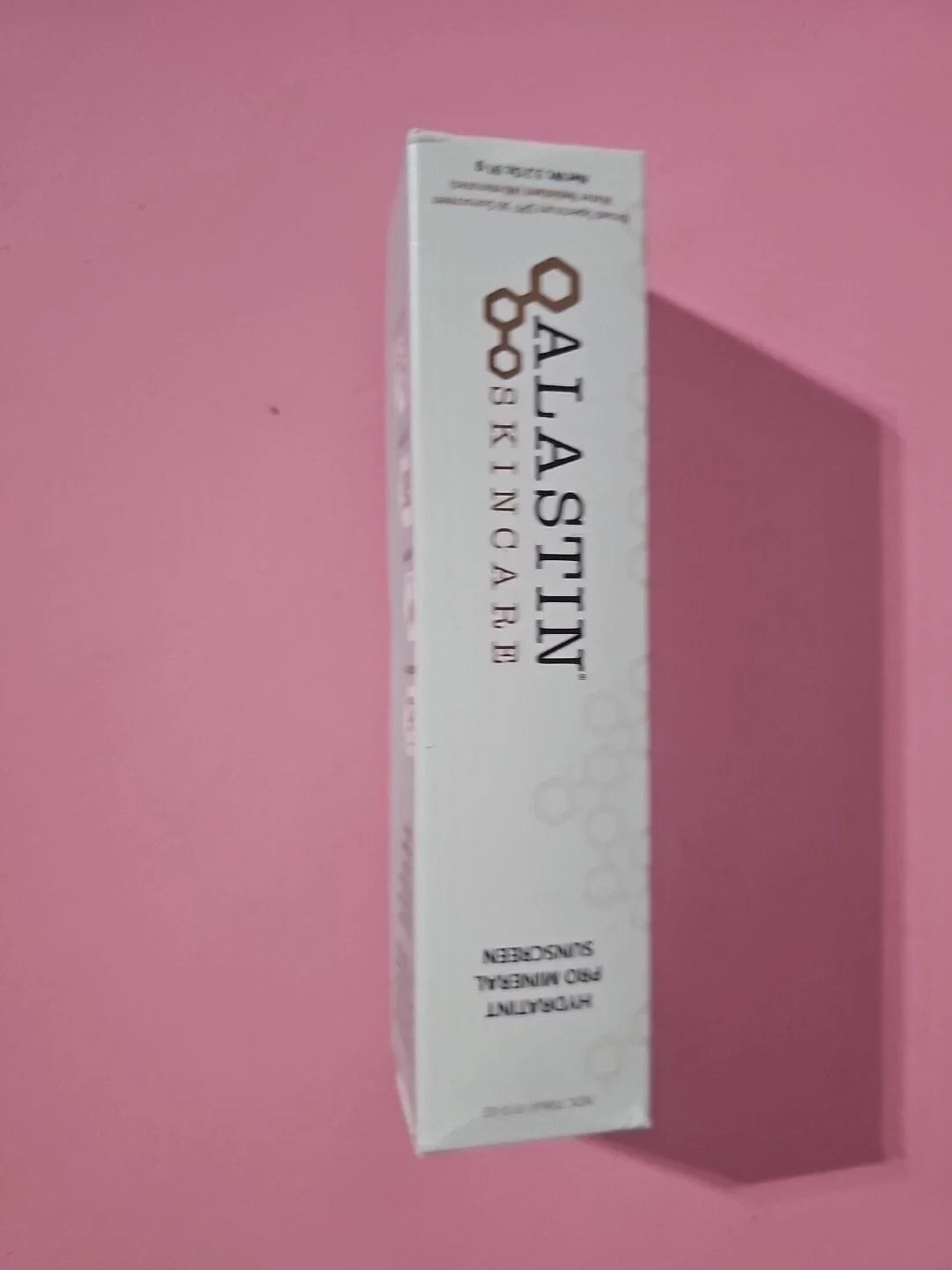 A white rectangular box of ALASTIN SKINCARE Hydrating Sunscreen, shown standing upright against a solid pink background with a soft shadow cast to the right.