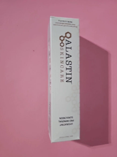 A white rectangular box of ALASTIN SKINCARE Hydrating Sunscreen, shown standing upright against a solid pink background with a soft shadow cast to the right.