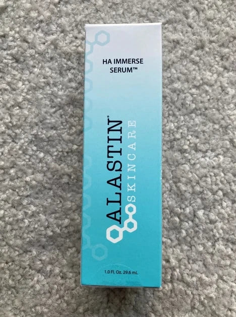 A tall rectangular box of ALASTIN SKINCARE HA IMMERSE SERUM in light blue and white, placed on a gray textured carpet background.