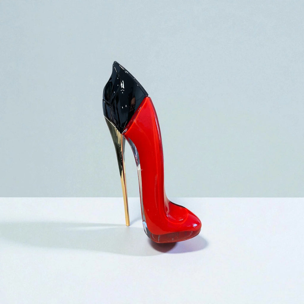 A glossy red high-heeled shoe sculpture or decorative object with a black pointed top and a slim metallic gold heel, standing on a white surface against a light gray background.