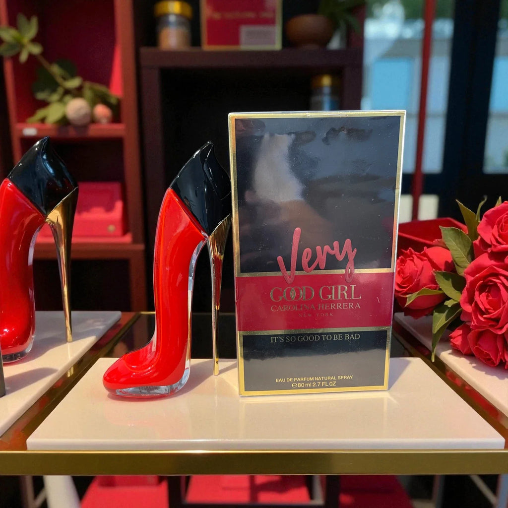 A boxed fragrance product labeled 'Very God Girl' by Carolina Herrera stands on a display table, flanked by two red-and-black high-heel-shaped objects and red roses in a retail-like setting.