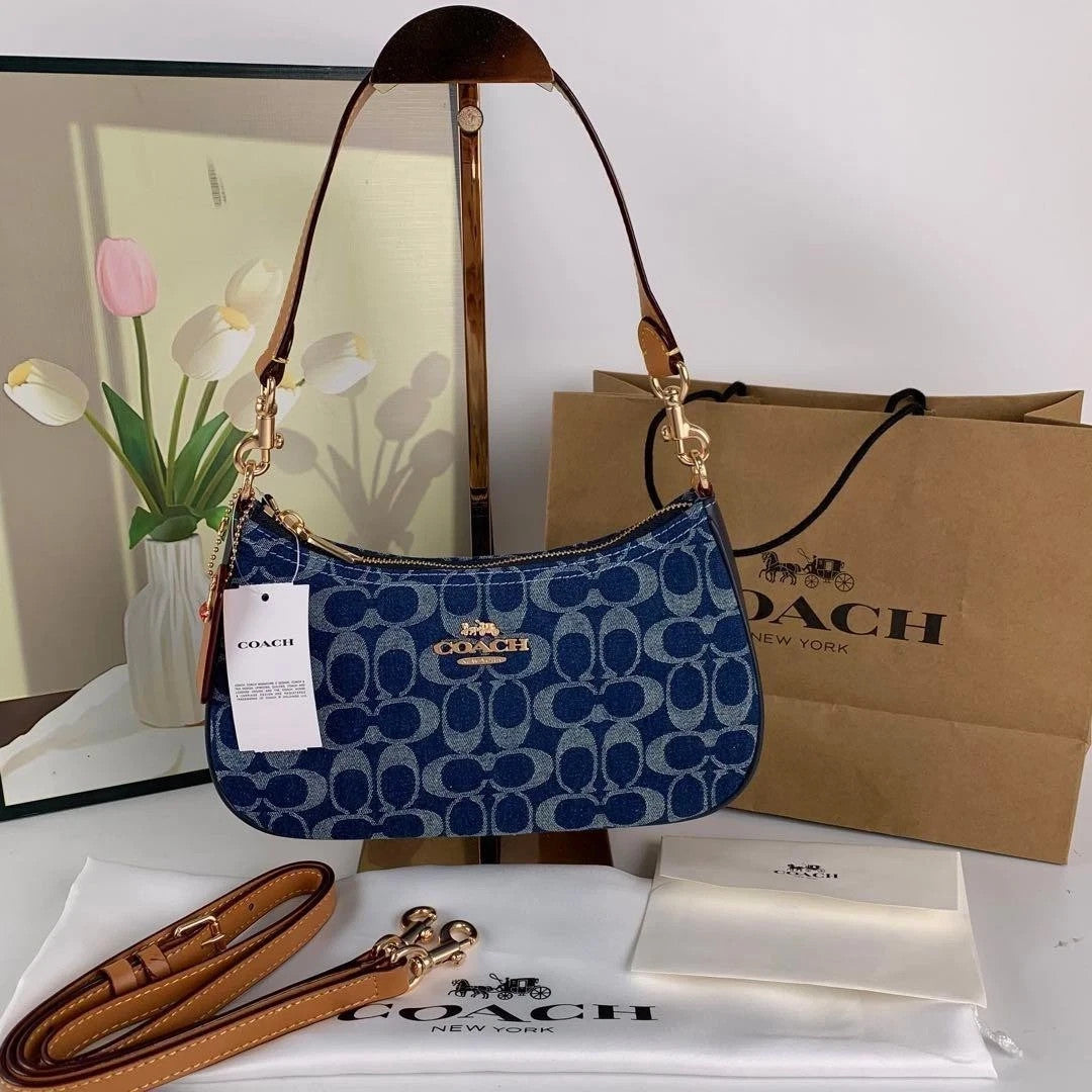 Blue Coach handbag with a matching strap on a white surface, with a Coach branded box and paper in the background.