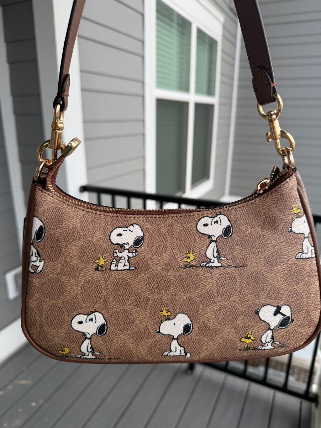 A brown shoulder bag featuring a repeating Snoopy and Woodstock print, shown hanging against a gray house exterior background.