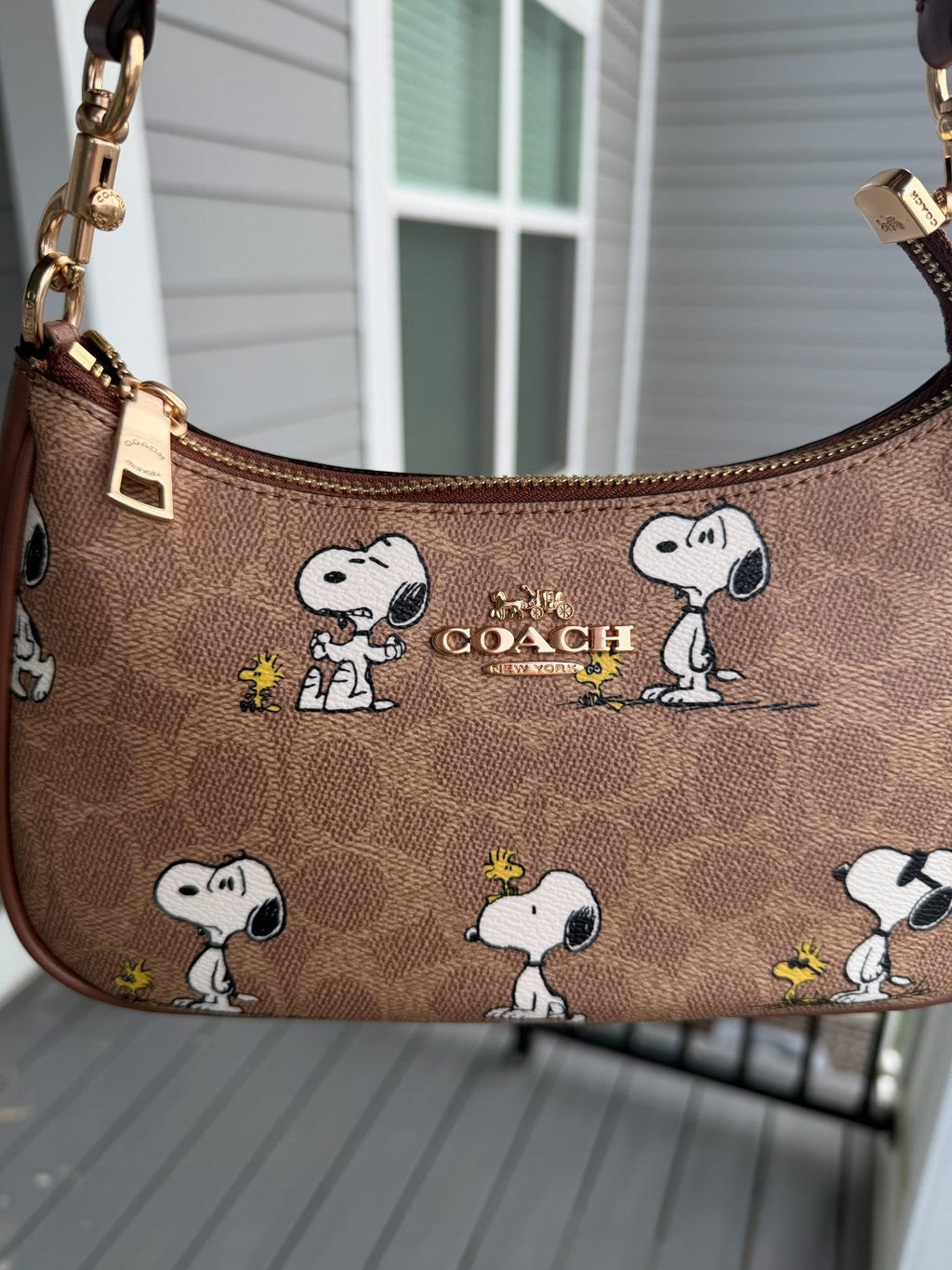A Coach shoulder bag featuring a brown signature canvas pattern with repeated Snoopy and Woodstock illustrations, gold-tone hardware, and the Coach logo on the front, photographed outdoors against a house wall and deck.