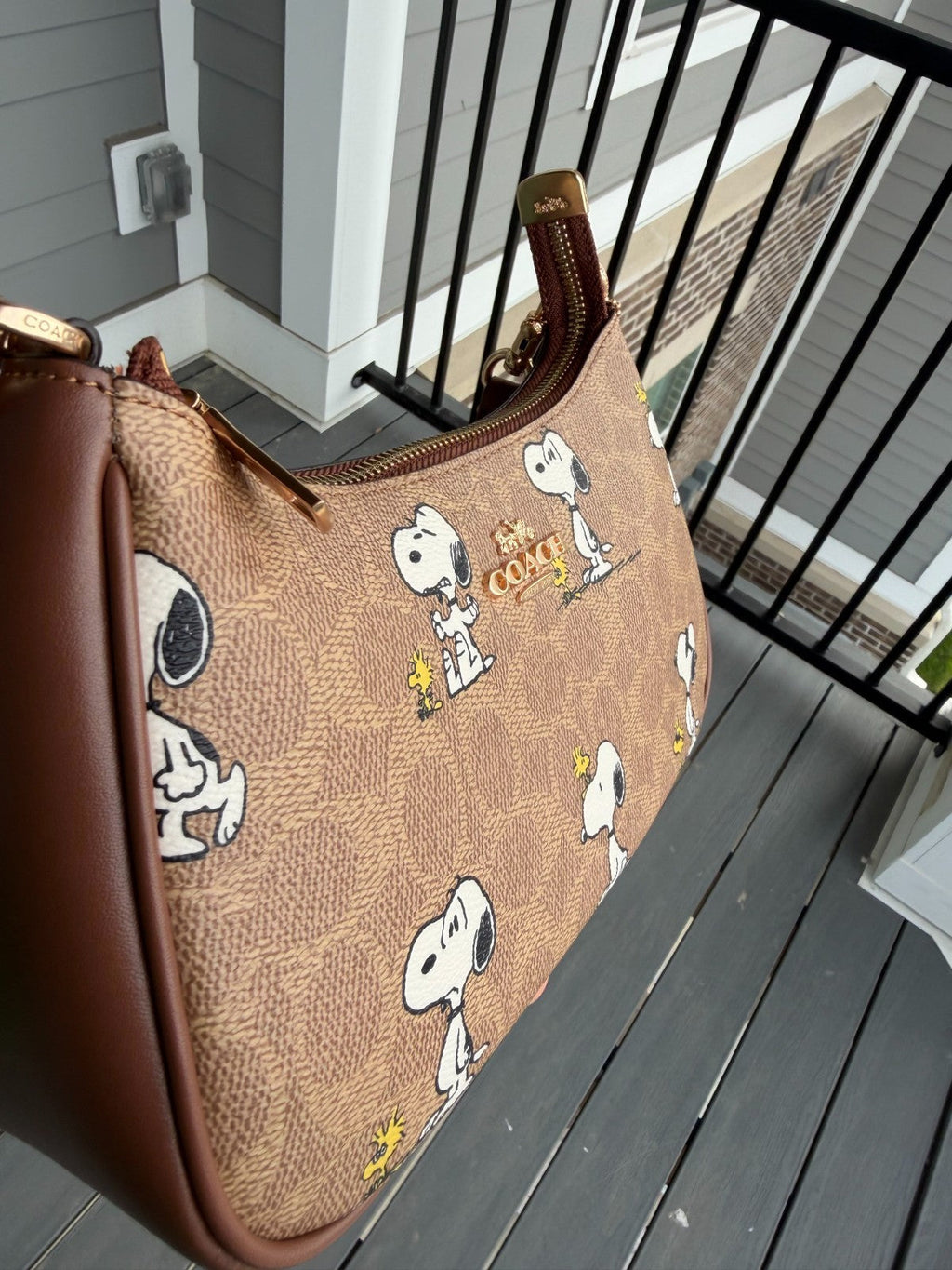 A Snoopy-themed shoulder bag with a brown monogram pattern, brown leather trim, and a gold-tone zipper, shown resting on a gray wooden deck next to a black metal railing.