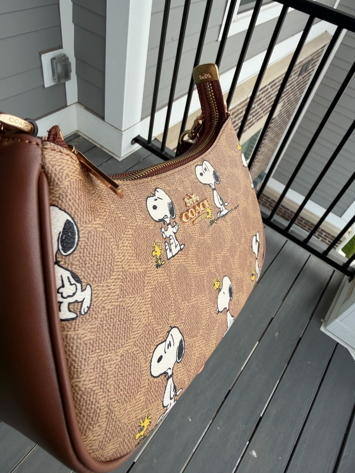 A Snoopy-themed shoulder bag with a brown monogram pattern, brown leather trim, and a gold-tone zipper, shown resting on a gray wooden deck next to a black metal railing.