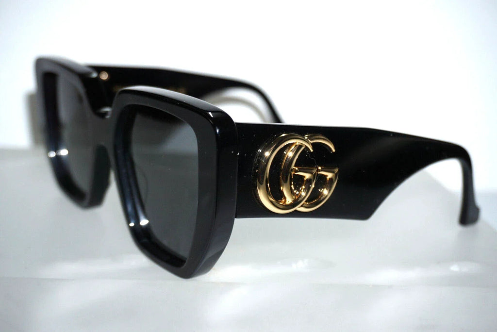 Black square sunglasses with a gold interlocking GG logo on the temple, displayed on a reflective white surface.