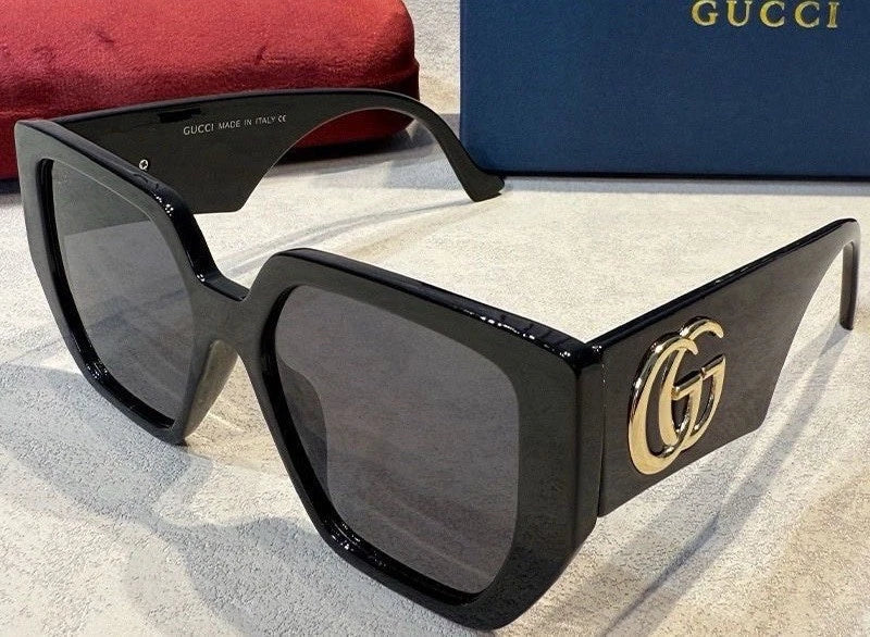 Gucci oversized square sunglasses with a glossy black frame, dark lenses, and a gold interlocking GG logo on the temple, shown with a red pouch and blue Gucci box in the background.