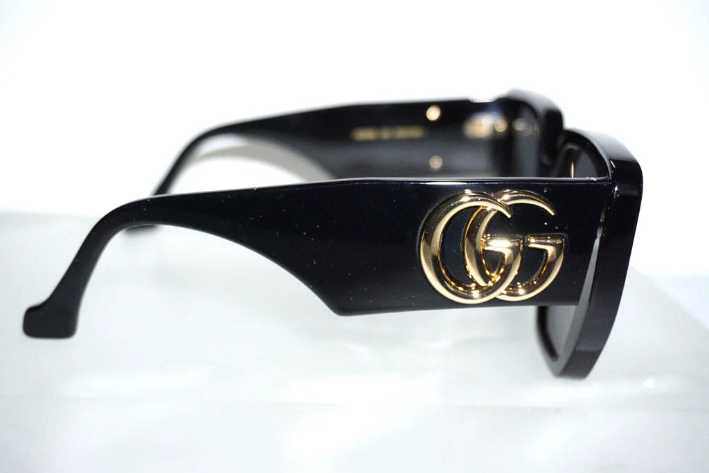 Black Gucci sunglasses with a gold interlocking GG logo on the temple, shown on a white reflective surface.