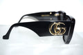 Black Gucci sunglasses with a gold interlocking GG logo on the temple, shown on a white reflective surface.