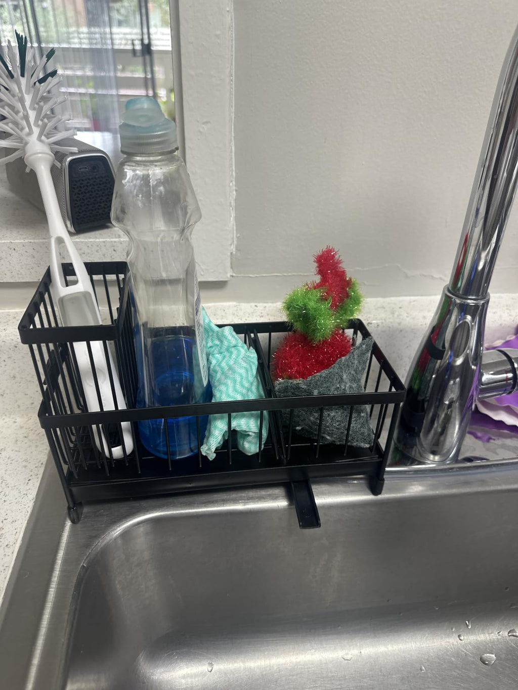 A black wire sink organizer basket hanging over a stainless steel sink, holding a white dish brush, a clear bottle, blue soap, a mint green sponge, and a red-and-green scrubber.