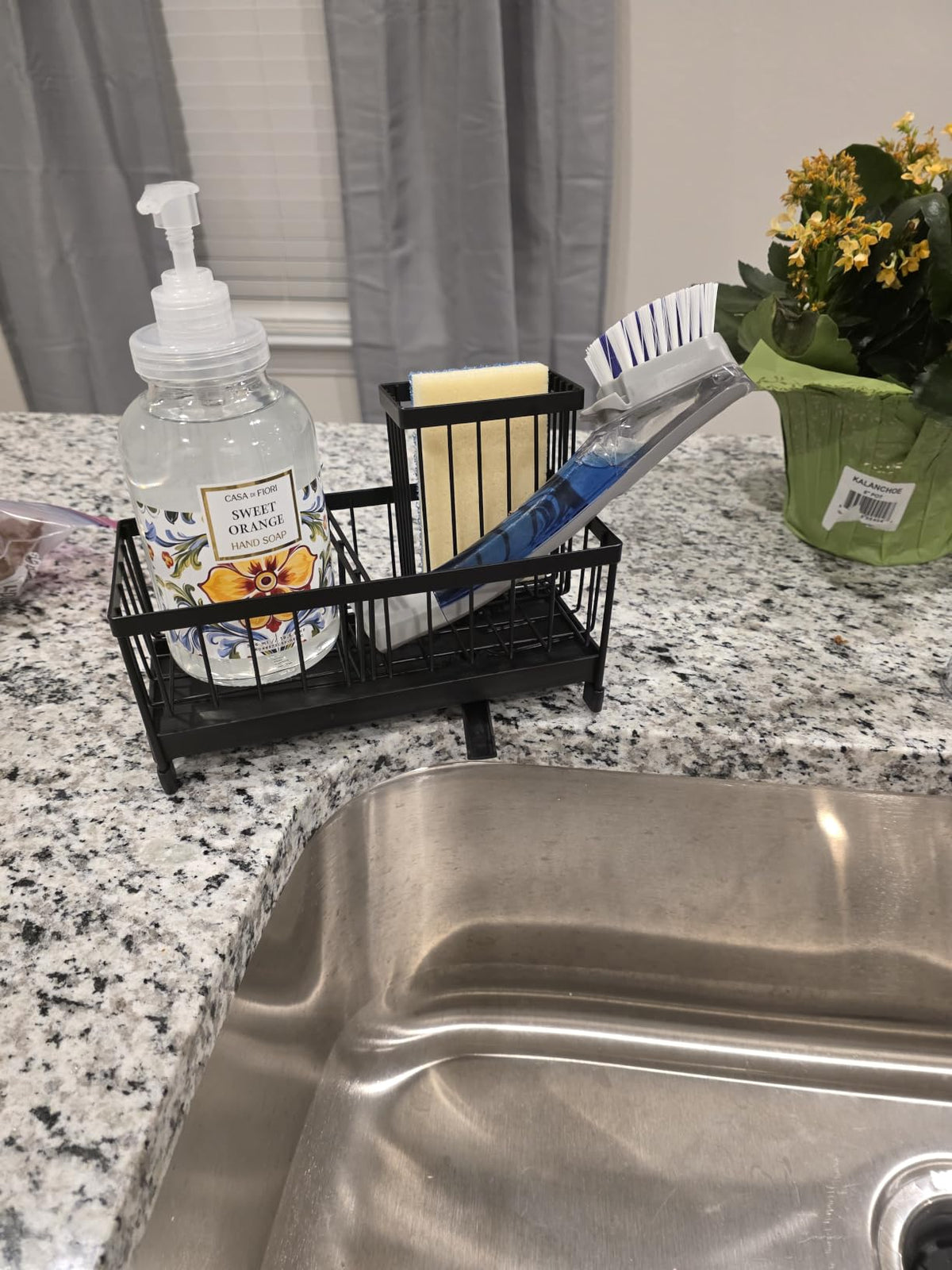A black wire sink organizer basket holding a sponge, toothbrush, and other small items, placed on a granite countertop next to a stainless steel sink, soap dispenser, and potted yellow flowers.