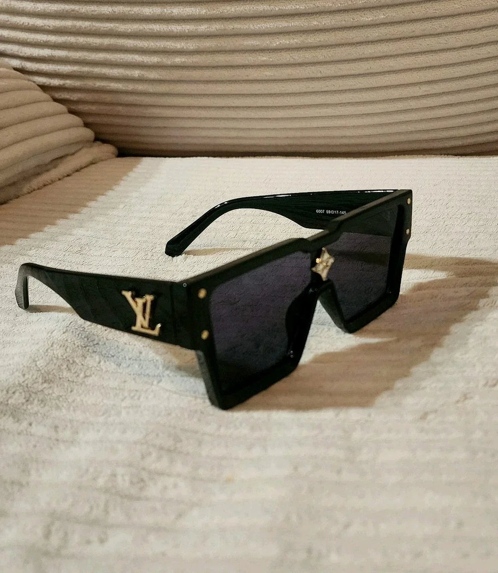 Louis Vuitton oversized square sunglasses with a glossy black frame, dark lenses, and gold-tone LV logos on the temples, resting on a textured beige fabric surface.