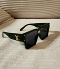 Louis Vuitton oversized square sunglasses with a glossy black frame, dark lenses, and gold-tone LV logos on the temples, resting on a textured beige fabric surface.
