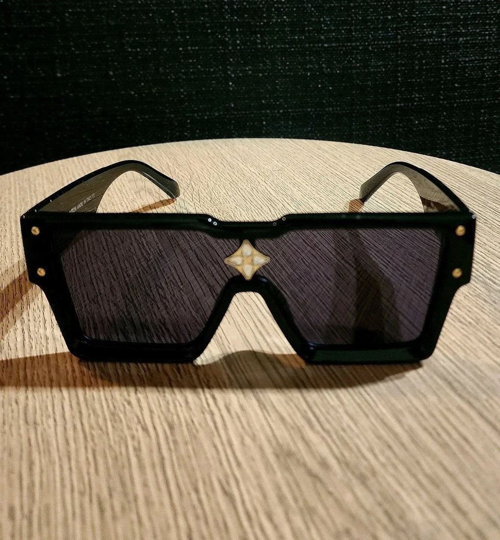 A pair of oversized square sunglasses with a glossy black frame, dark tinted lenses, and a small white geometric logo centered above the lenses, resting on a light wood surface.