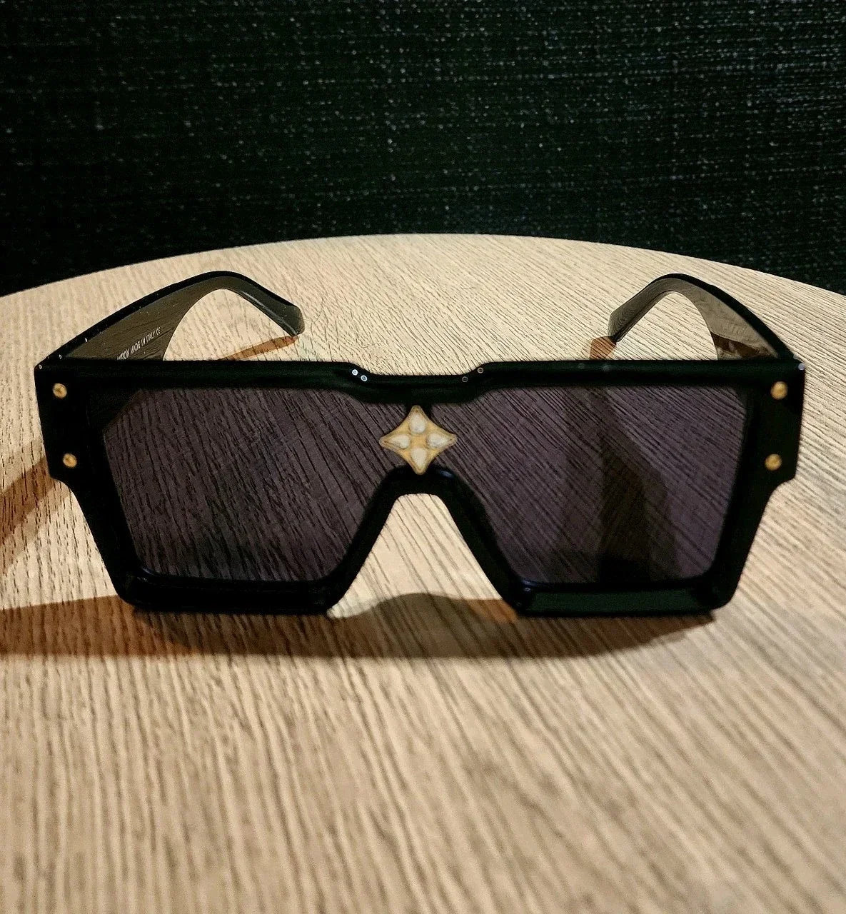 A pair of oversized square sunglasses with a glossy black frame, dark tinted lenses, and a small white geometric logo centered above the lenses, resting on a light wood surface.