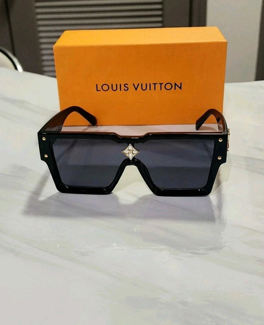 Louis Vuitton oversized square sunglasses with dark lenses and a small star detail on the bridge, displayed in front of an orange Louis Vuitton box on a light marble surface.