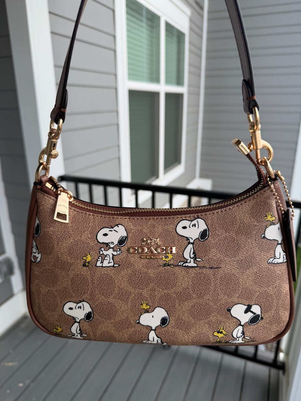 A brown Coach shoulder bag featuring an all-over Snoopy and Woodstock pattern, hanging on a gray porch in front of a house window.