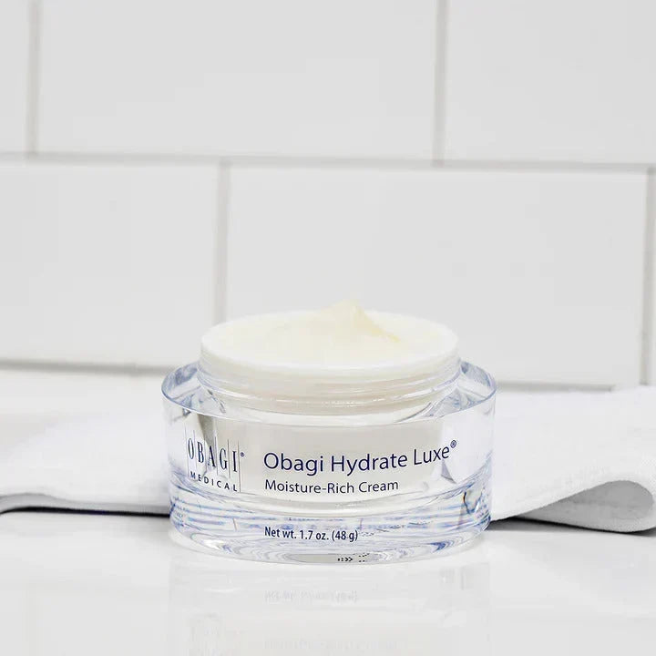 A clear plastic jar of Obagi Magical Obagi Hydrate Luxe Moisture-Rich Cream with white cream inside, set on a white surface with a white tiled background.
