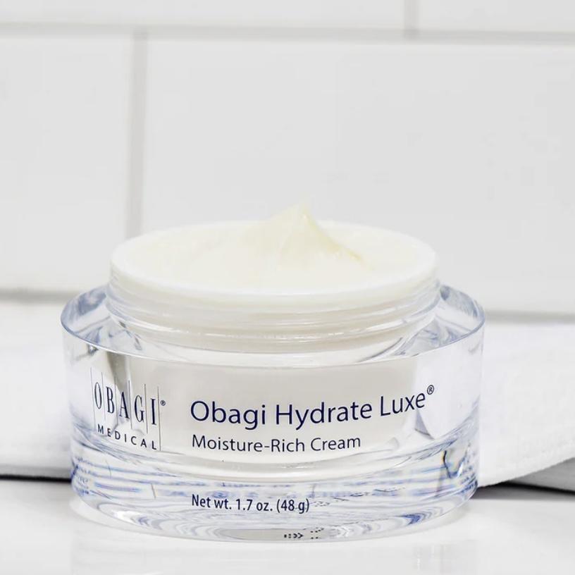 A clear cosmetic jar of Obagi Hydrate Luxe Moisture-Rich Cream with white cream visible on top, set against a clean white tiled background.