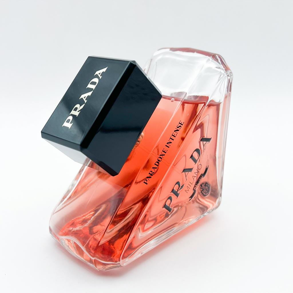 Prada Parfum Intense perfume bottle with a faceted clear glass body filled with vibrant orange-red liquid, a black cap, and silver Prada branding.
