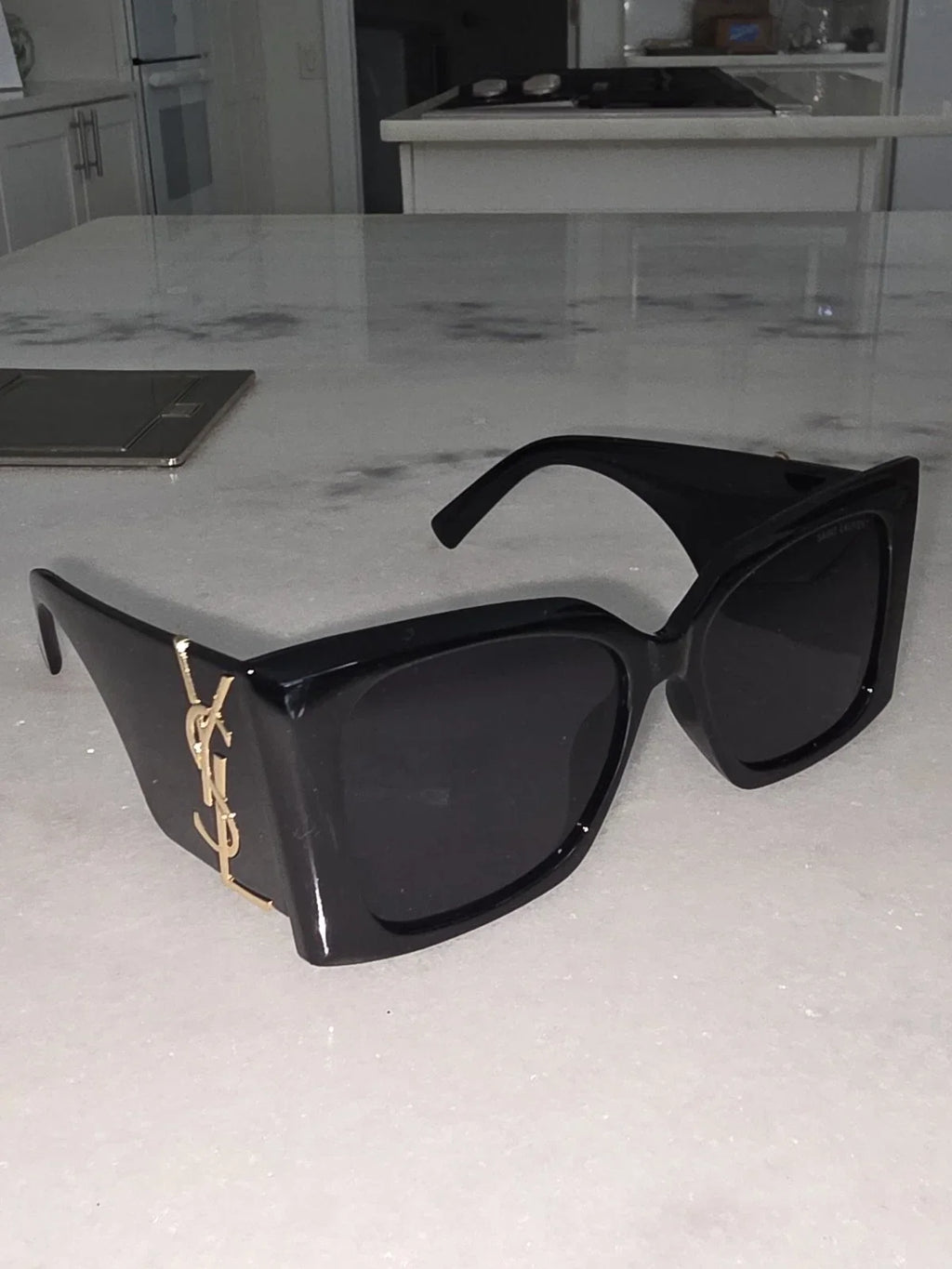 Black oversized square sunglasses with dark lenses and a gold YSL logo on the temple, resting on a white marble kitchen countertop.