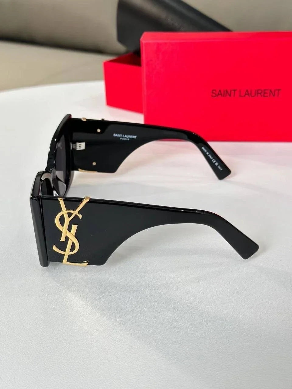 Saint Laurent square-frame sunglasses in glossy black with a gold YSL logo on the temple, shown with a red Saint Laurent box in the background.