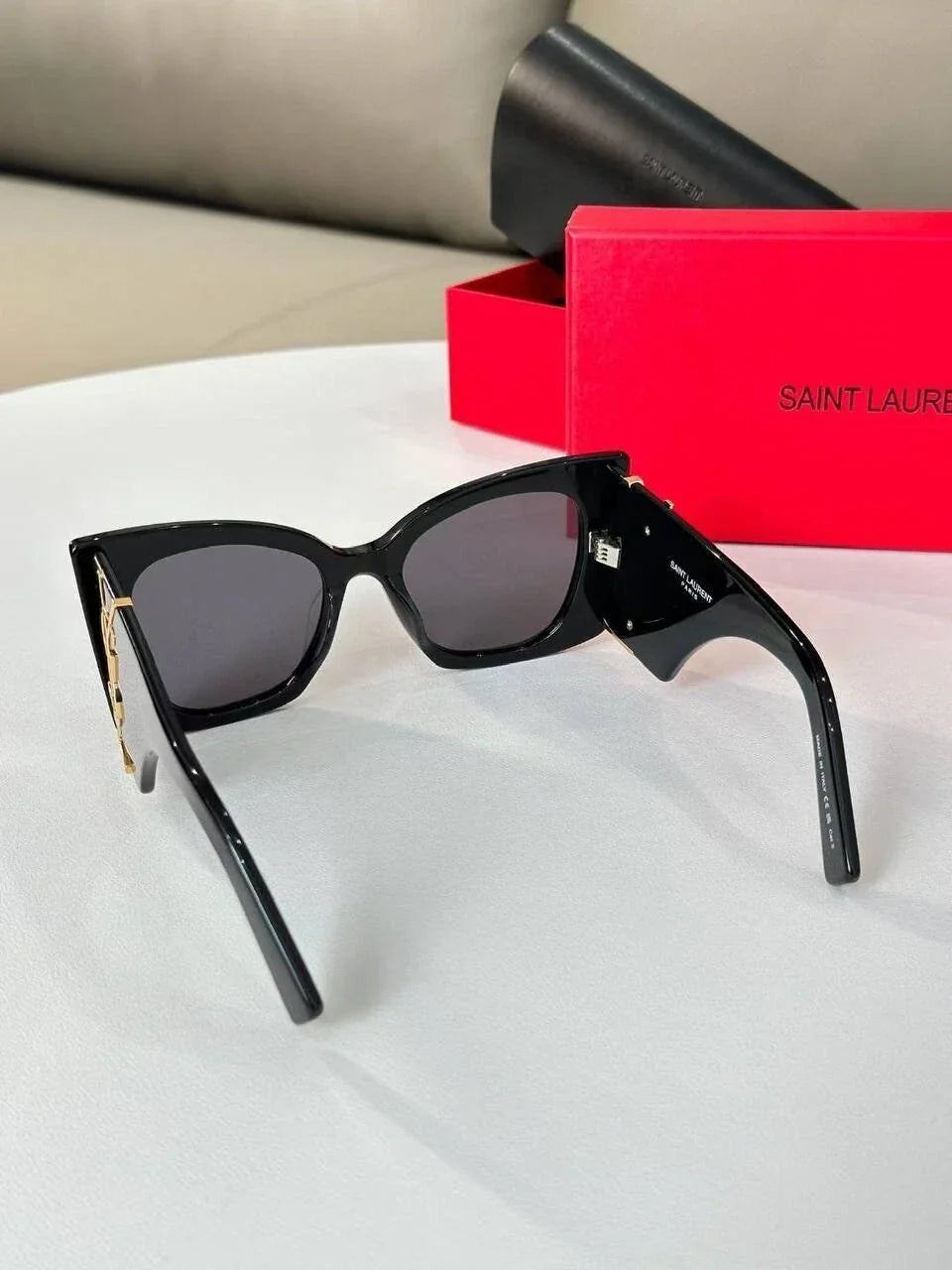 Saint Laurent sunglasses with black cat-eye frames, dark lenses, and gold-tone hardware, displayed next to a red Saint Laurent box on a white surface.