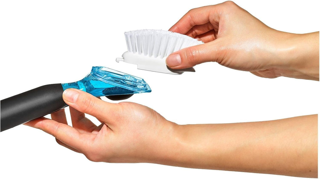 A close-up of a person's hands demonstrating a cleaning attachment. One hand holds a black handheld device with a clear blue water reservoir, while the other hand holds a white bristle brush against the nozzle opening to show how the brush is inserted or used.