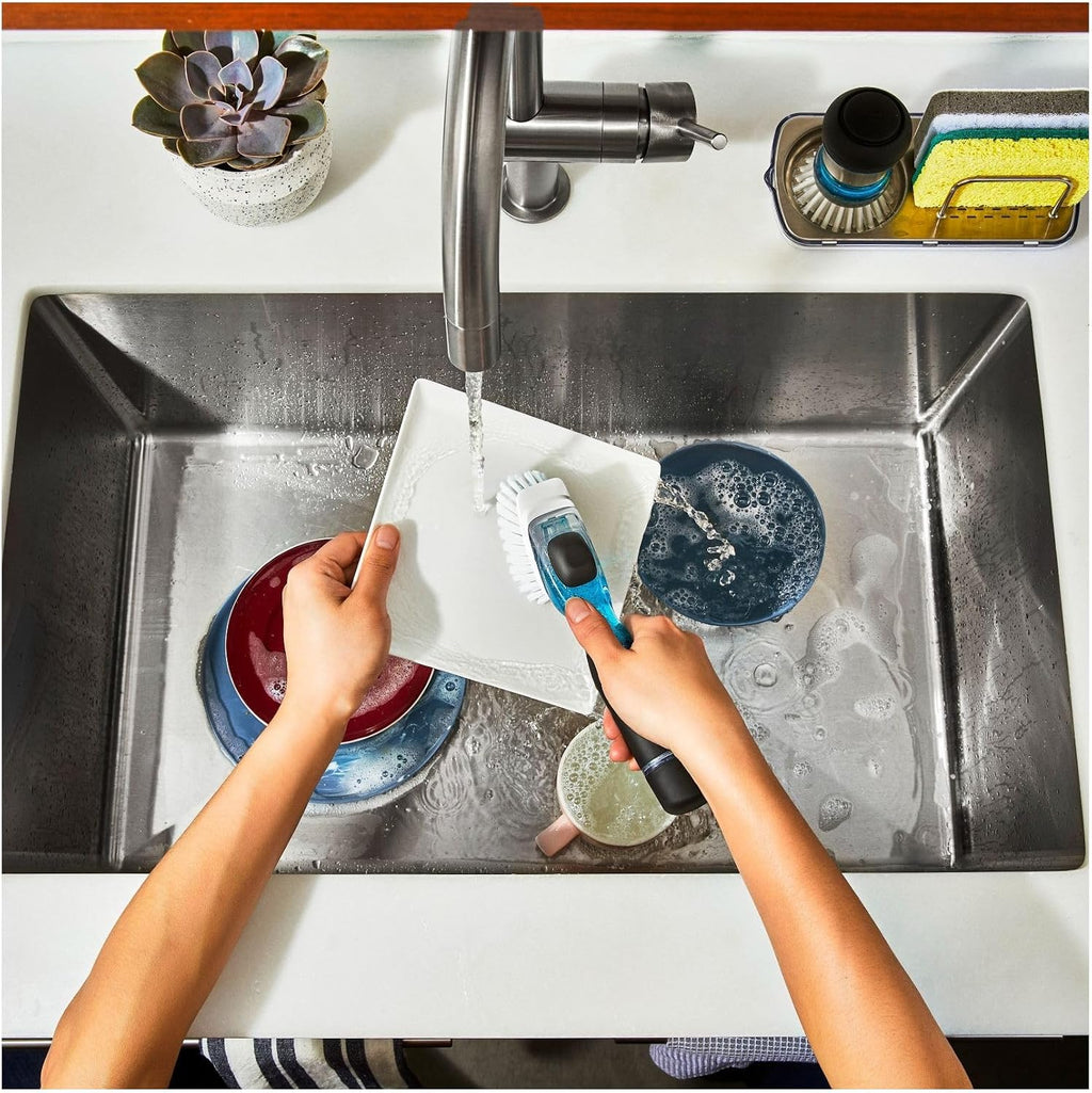 Top-down view of a person washing a white plate in a stainless steel kitchen sink, using a blue and black handheld scrubber brush. Dish soap, sponges, and other dishes are visible in the sink, with a faucet and countertop around it.
