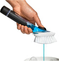 A hand holding a handheld cleaning brush with a black handle, blue transparent body, white bristles, and a turquoise cord above a sink with ice on a white background.