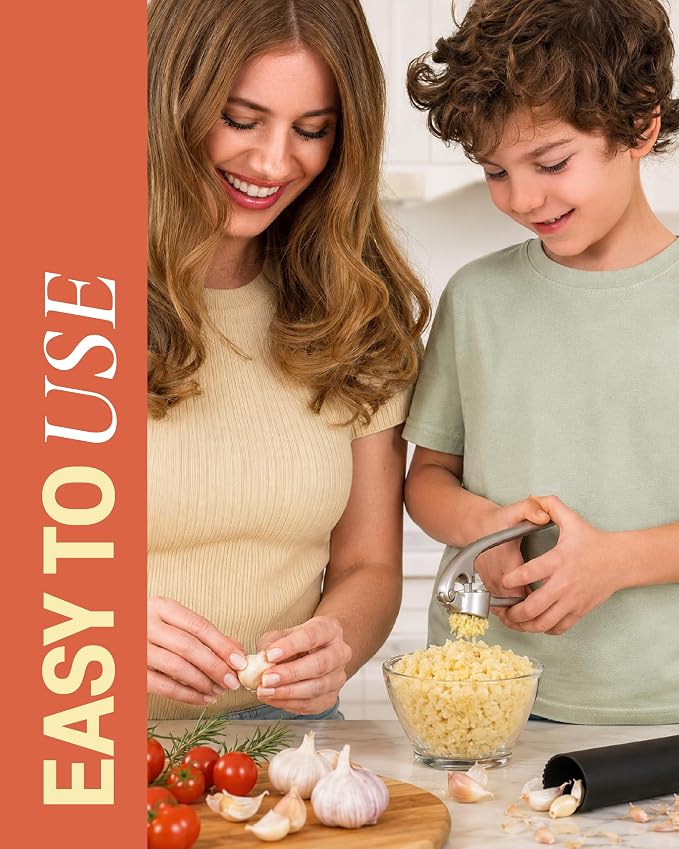 A woman and a boy demonstrate a garlic press in a bright kitchen setting. The boy is actively using the silver garlic press to extract garlic from a bowl of peeled corn kernels, while the woman stands beside him.