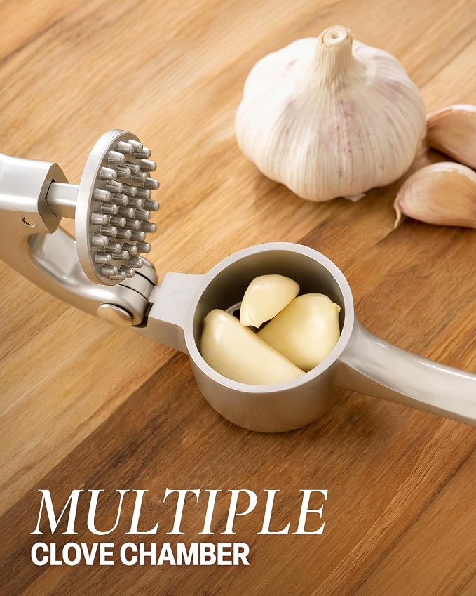 A stainless steel garlic press with a textured clove chamber and handle, shown with garlic cloves inside and whole garlic bulbs on a wooden surface. Text overlay reads 'MULTIPLE CLOVE CHAMBER'.