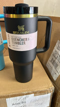 A black Stanley Quencher Hot Tumbler with a gold band and a pink-and-white label, shown standing on a cardboard box in a warehouse-like setting.