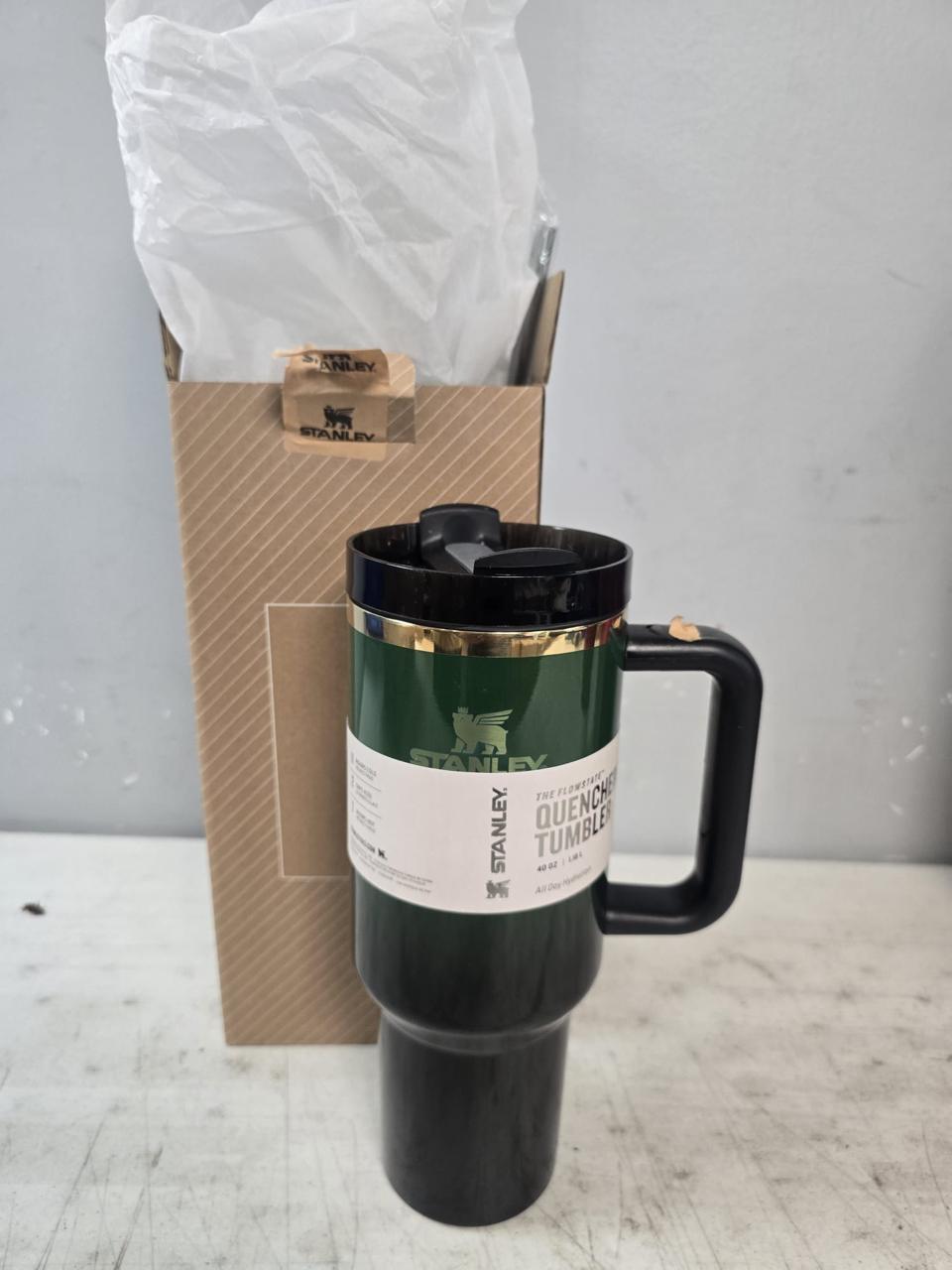 A Stanley Ogunmian Tumbler in black with a dark green band and a white label, shown next to its brown cardboard packaging box with a clear plastic bag on top, on a light gray surface.