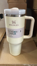 A Stanley QuenchState Quencher H2.0 travel tumbler in cream with a white lid and handle, shown on a cardboard surface with a brown box in the background.