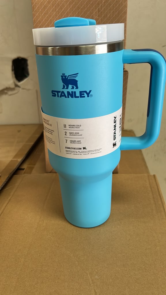 A light blue Stanley travel mug with a matching blue handle and lid, featuring a white paper label on the front and a stainless steel rim, set against a cardboard background.