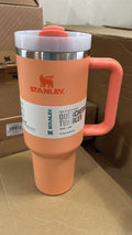 A Stanley QuenchMaster travel tumbler in a peach-orange color, featuring a white lid with a coral accent, a stainless steel rim, and a white product label on a cardboard background.