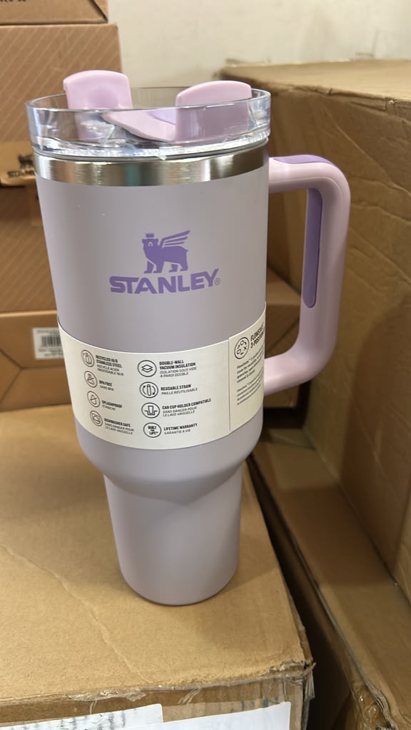 A lavender Stanley travel tumbler with a matching lavender handle and lid, sitting on a cardboard shipping box in a warehouse setting.