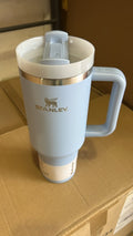 A light blue Stanley travel mug with a white lid and handle, resting on a brown cardboard box.