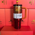 Stanley Quencher Tumbler in red and gold, featuring a built-in handle and straw, displayed in front of red Stanley packaging boxes.