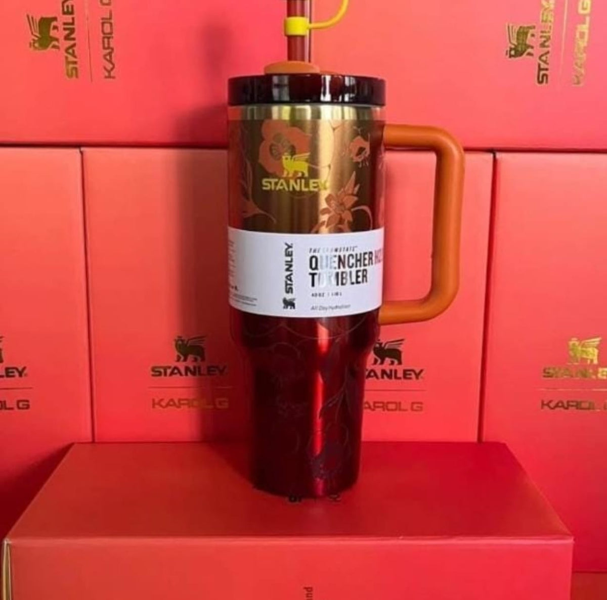 Stanley Quencher Tumbler in red and gold, featuring a built-in handle and straw, displayed in front of red Stanley packaging boxes.