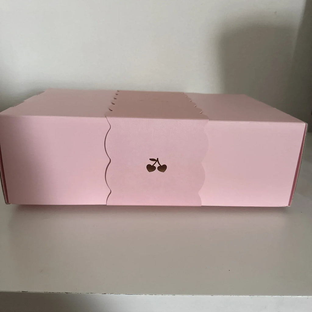 A pink rectangular product box with a scalloped front flap and a small cherry graphic, photographed on a light surface against a plain wall.
