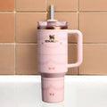 A pink Stanley travel mug with a handle, a clear lid with a white straw, and rose gold trim, set against a background of tan ceramic tiles.